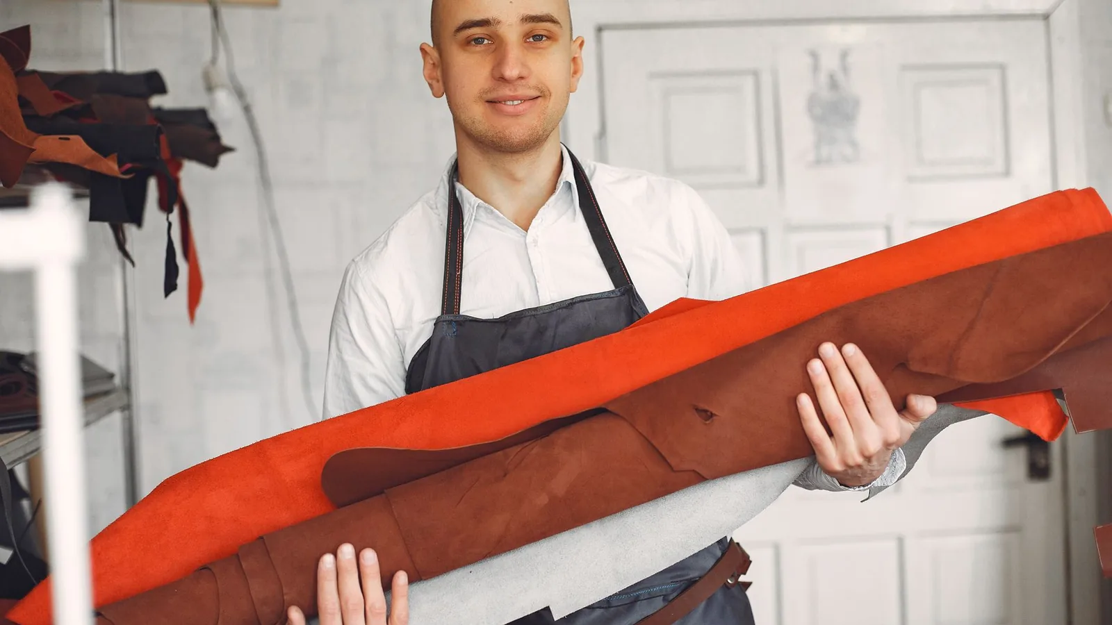 Positive adult male artisan in formal shirt and apron holding velour cloth rolls while working in workshop