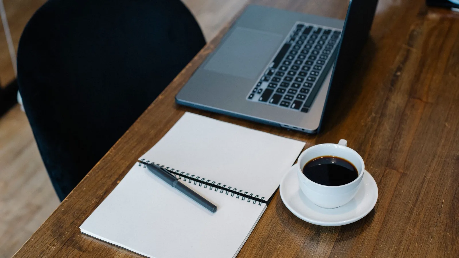 High angle of opened laptop and spiral notebook with blank pages placed on wooden table with cup of coffee and pen during workday