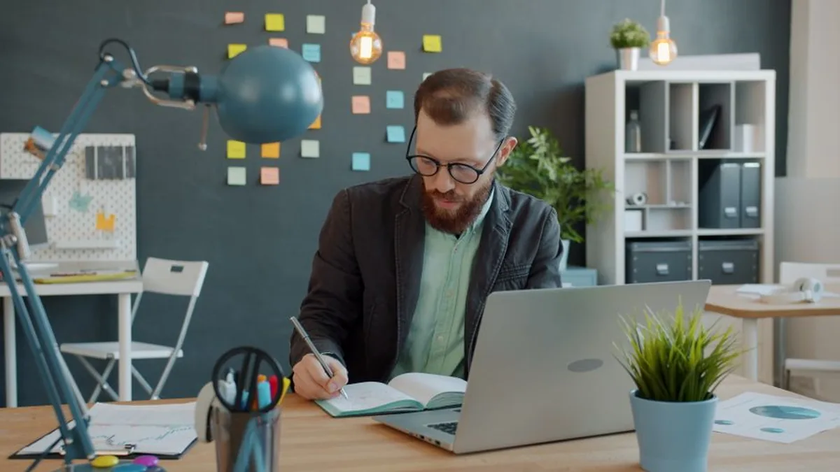 Business professional working at a desk with a laptop, taking notes in a creative office space.